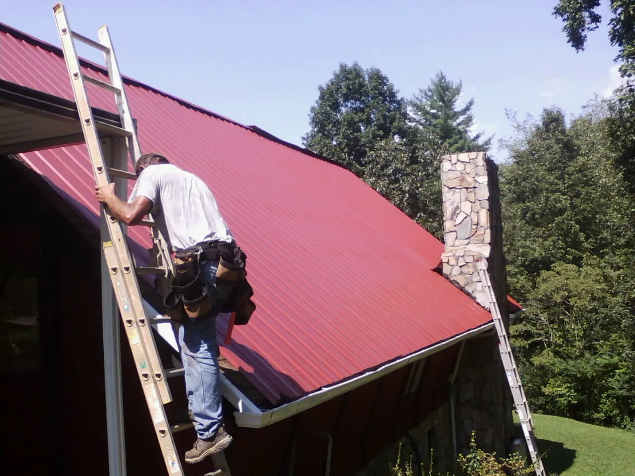 Roofer on a ladder beside a red metal roof and stone chimney — Soffit & Fascia Repair by Roofing Brothers in Shinnston